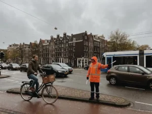Verkeersregelaar in oranje hesje dirigeert verkeer op Amsterdams kruispunt met traditionele grachtenpanden op achtergrond