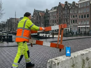 Verkeersregelaar in hesje past oranje-witte wegafzetting aan op Amsterdamse straat met gracht en bakstenen gebouwen