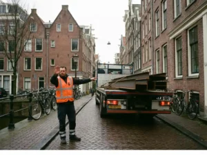 Nederlandse verkeersregelaar in oranje hesje dirigeert vrachtwagen met staalbalken op Amsterdamse straat met rode bakstenen