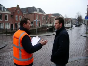 Nederlandse verkeersregelaar in oranje veiligheidsvest overlegt met projectmanager bij kruispunt met rode bakstenen gebouwen