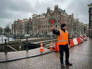 Verkeersregelaar in oranje vest met rood-groen bord op Amsterdamse gracht bij bouwwerkzaamheden aan kanaalburg